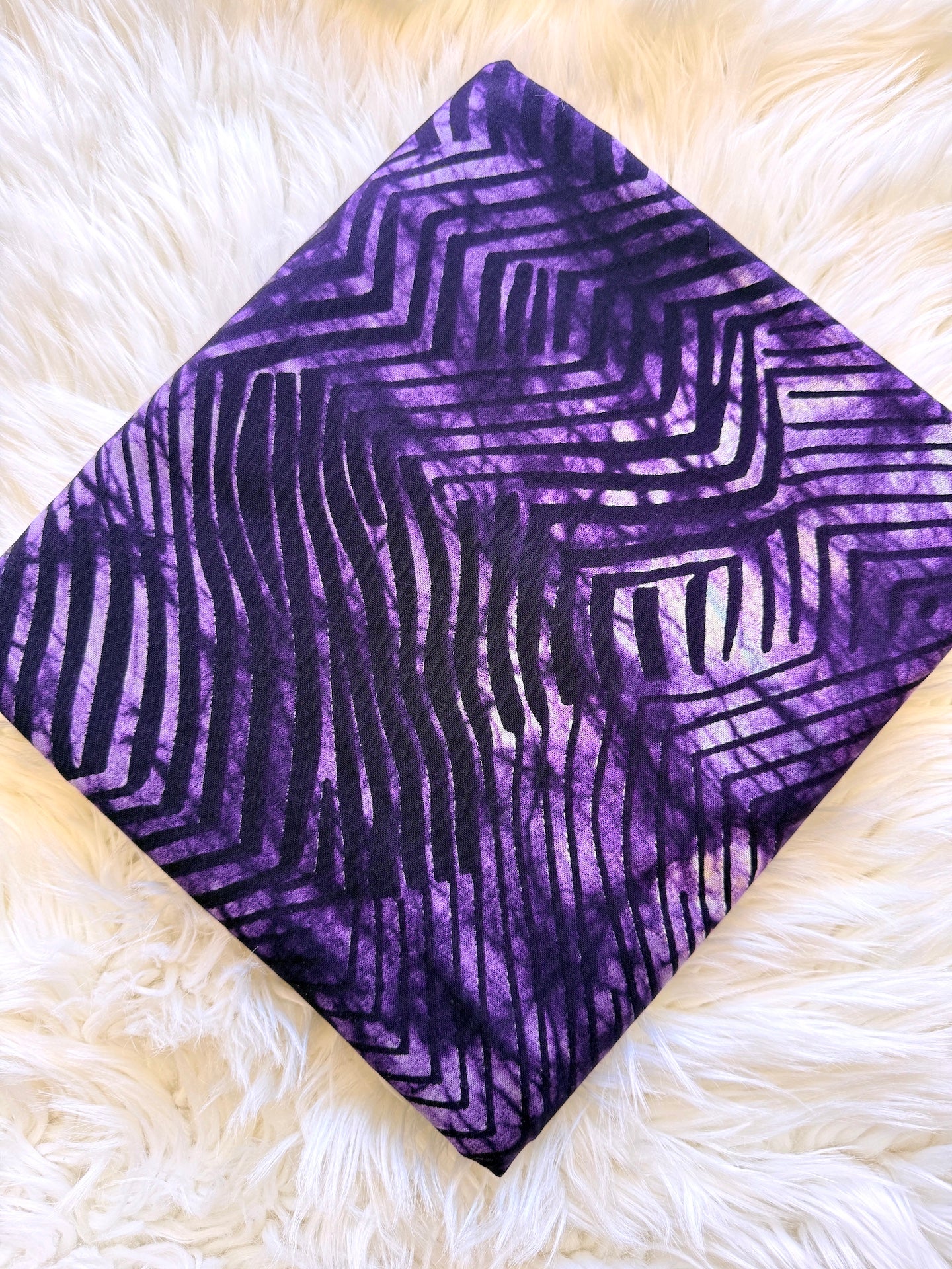A square fabric bandana or scarf with a purple and black geometric wavy pattern, laid flat on a white faux fur surface.