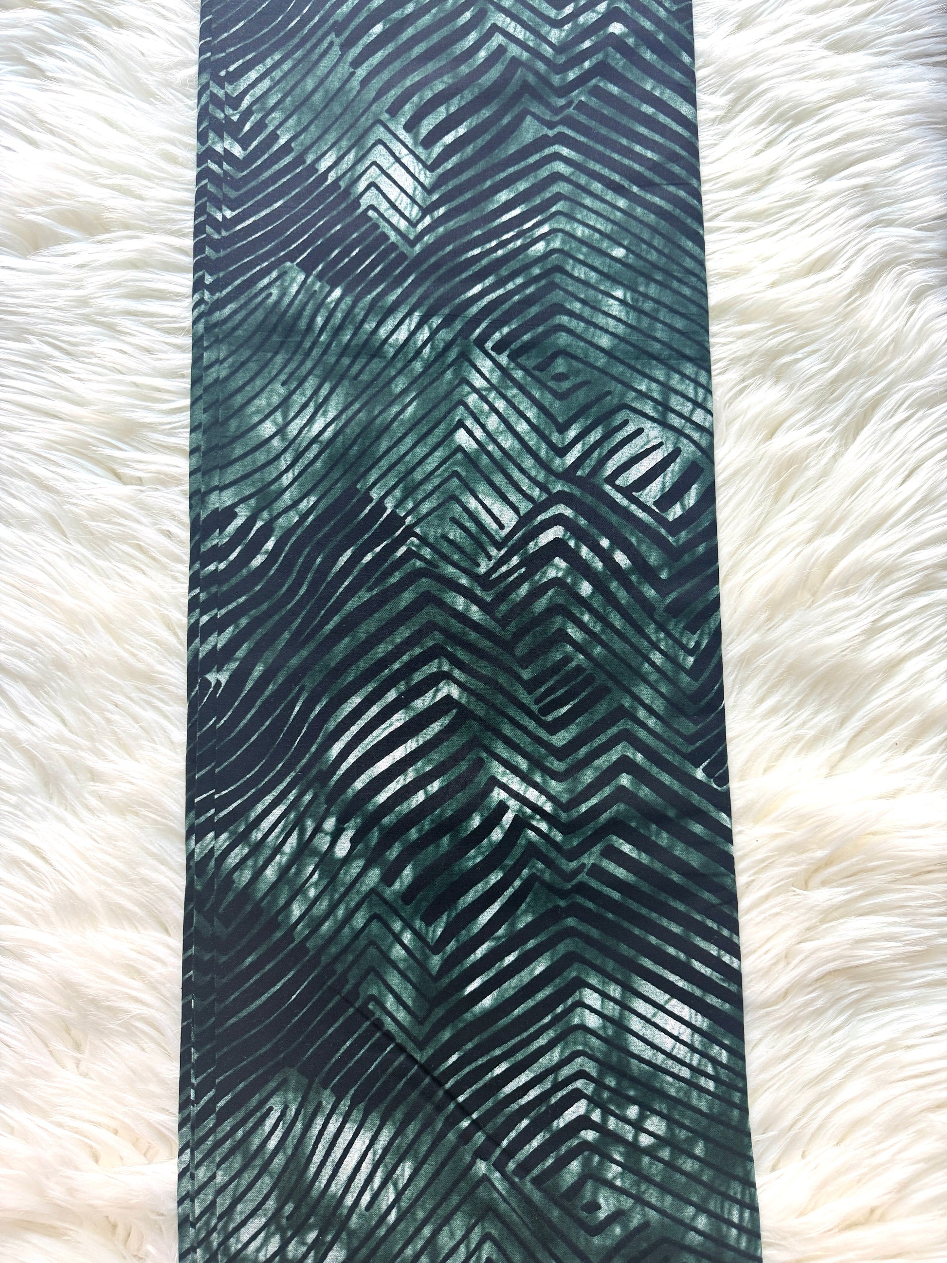 A long rectangular textile item with an abstract wavy chevron pattern in dark teal, green, and black, laid flat on a white faux-fur surface.