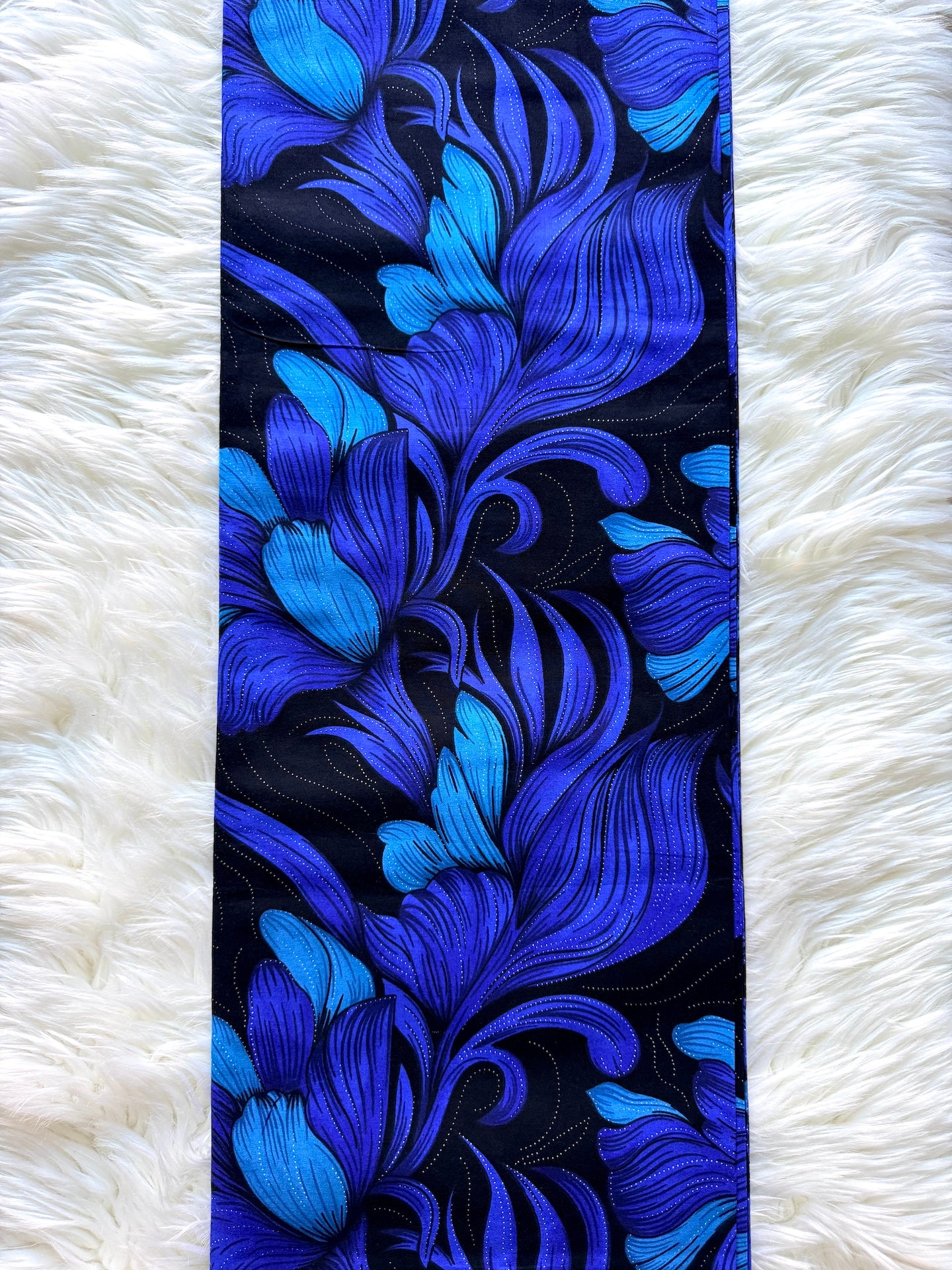 A long rectangular textile with an ornate floral or scroll-like pattern in vivid blue and cobalt tones on a black background, laid flat on a white faux-fur surface.