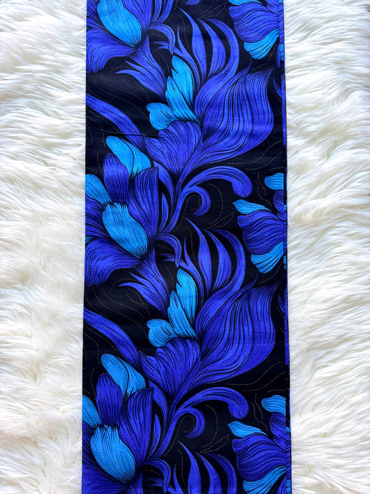A long rectangular textile with an ornate floral or scroll-like pattern in vivid blue and cobalt tones on a black background, laid flat on a white faux-fur surface.