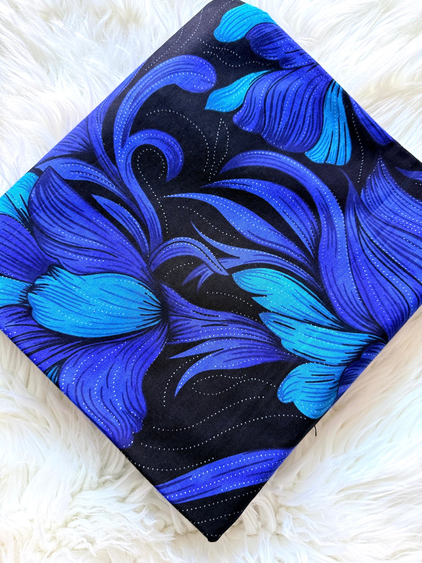 A square fabric item, likely a napkin or small scarf, featuring a bold abstract floral pattern in shades of blue and black, laid flat on a white faux fur surface.