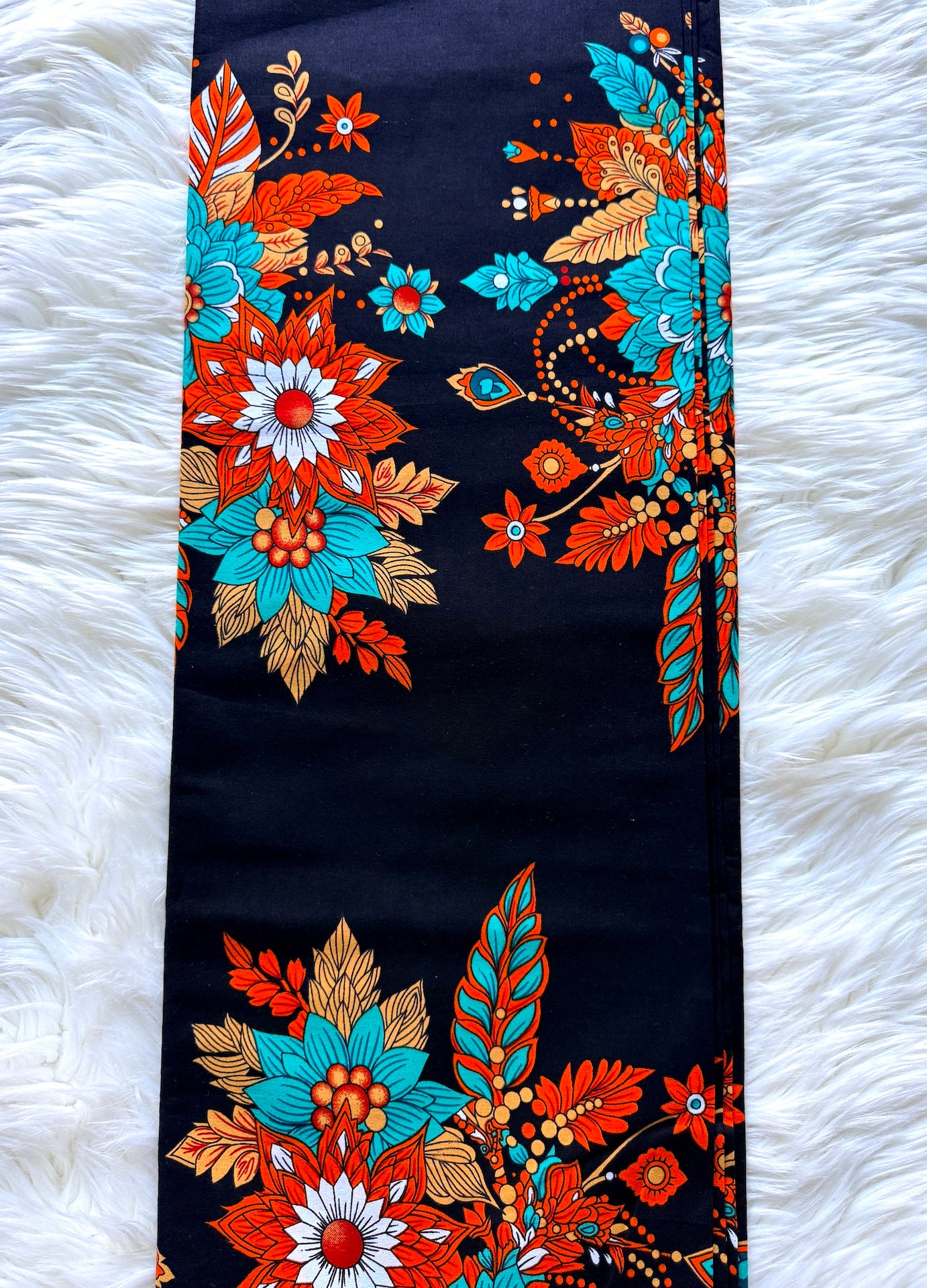 A long rectangular textile laid flat on a white faux-fur surface, featuring a bold floral and ornamental print in turquoise, rust orange, tan, white, and red on a very dark navy or near-black background.