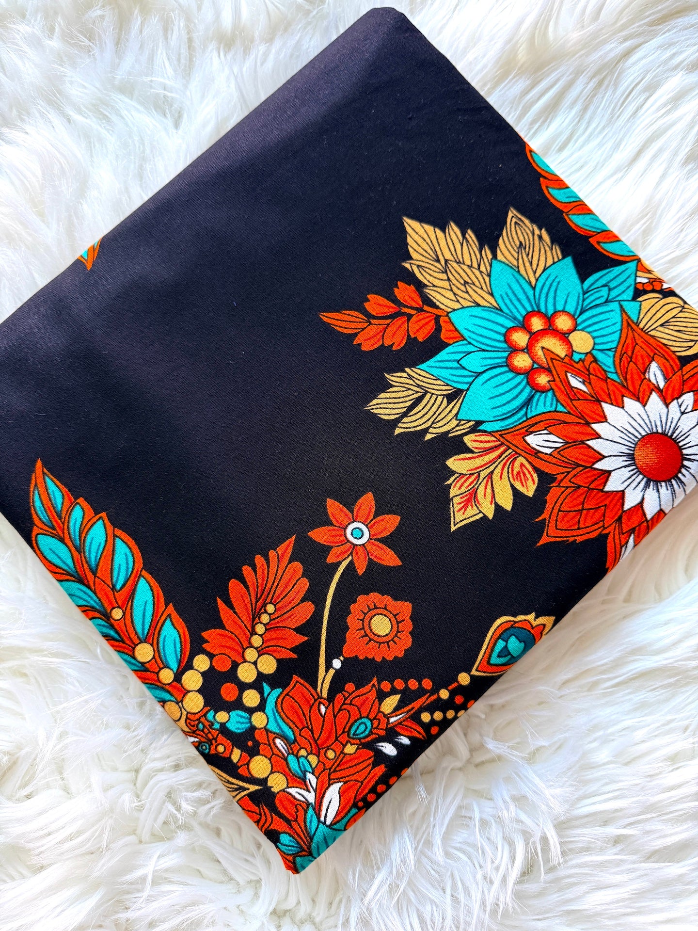 A folded black fabric item, likely a scarf or bandana, featuring a vibrant retro floral pattern in orange, turquoise, gold, and white, displayed on a white faux fur surface.
