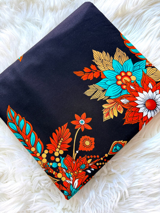 A folded black fabric item, likely a scarf or bandana, featuring a vibrant retro floral pattern in orange, turquoise, gold, and white, displayed on a white faux fur surface.
