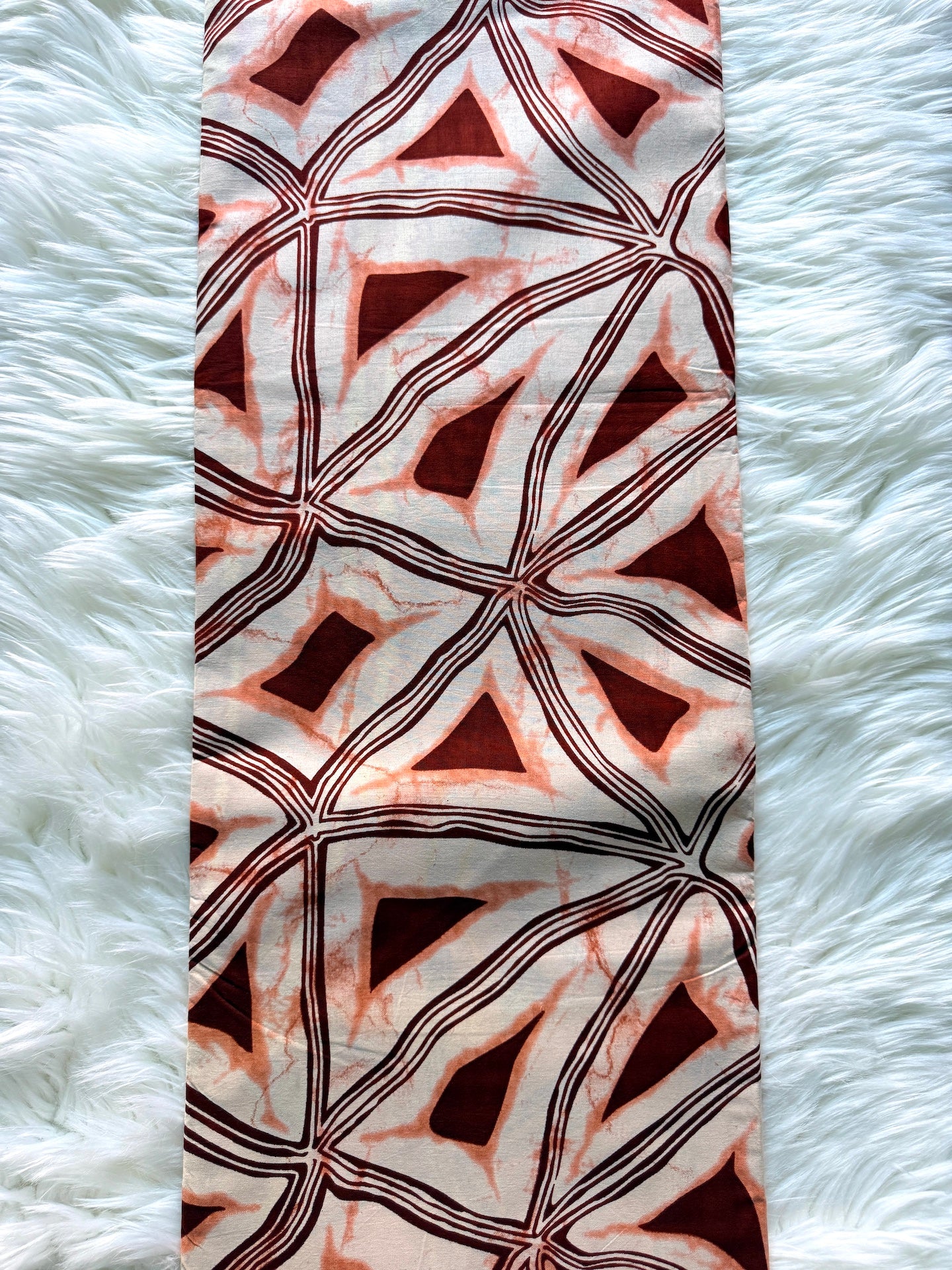 A vertical rectangular scarf or fabric panel featuring an abstract geometric pattern of triangles and diamonds in deep red, terracotta, and black over a warm off-white background, laid flat on a white faux fur surface.