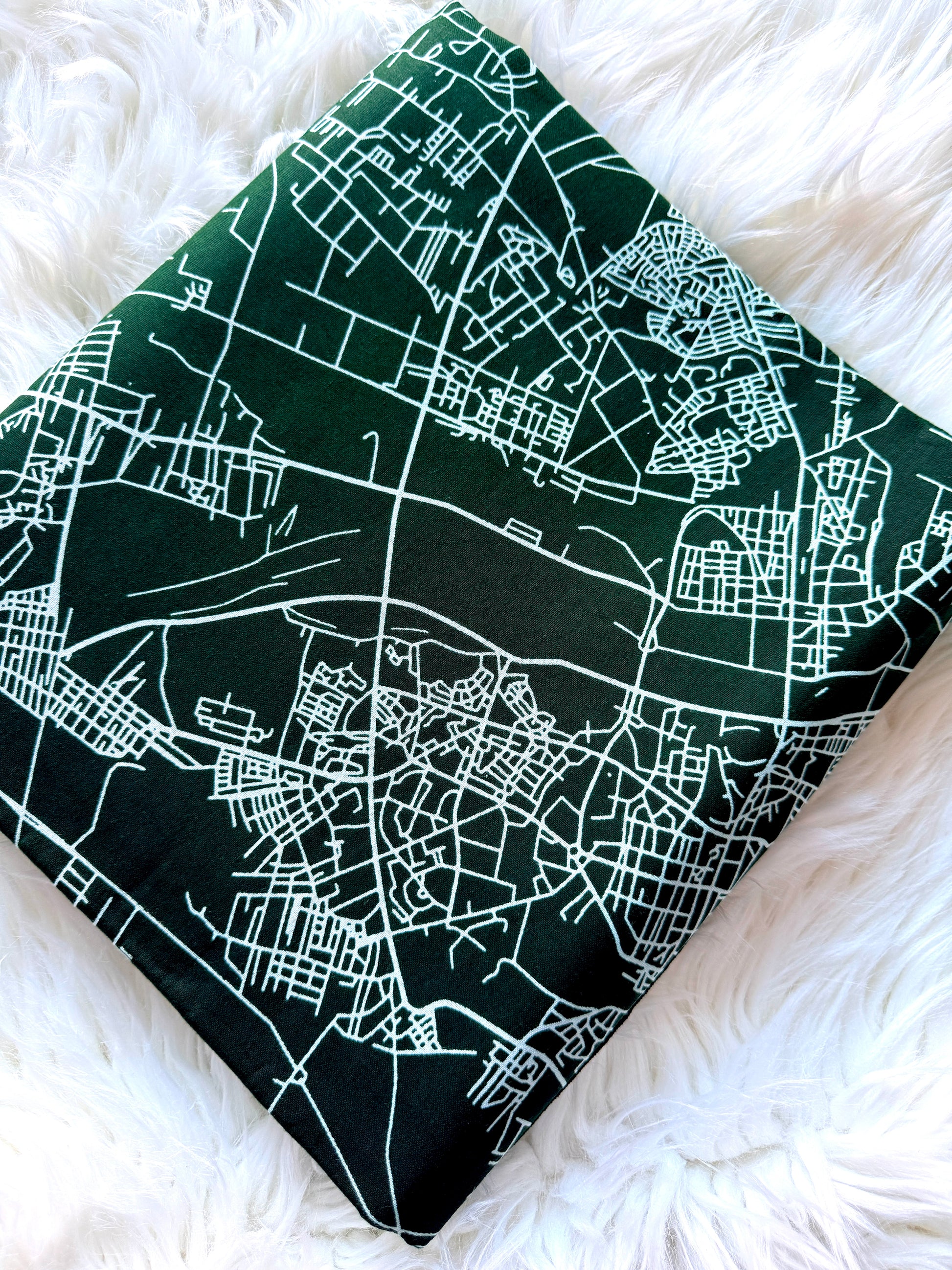 A square fabric pillow or cushion cover featuring a white street map print on a dark green background, displayed on a white faux fur surface.