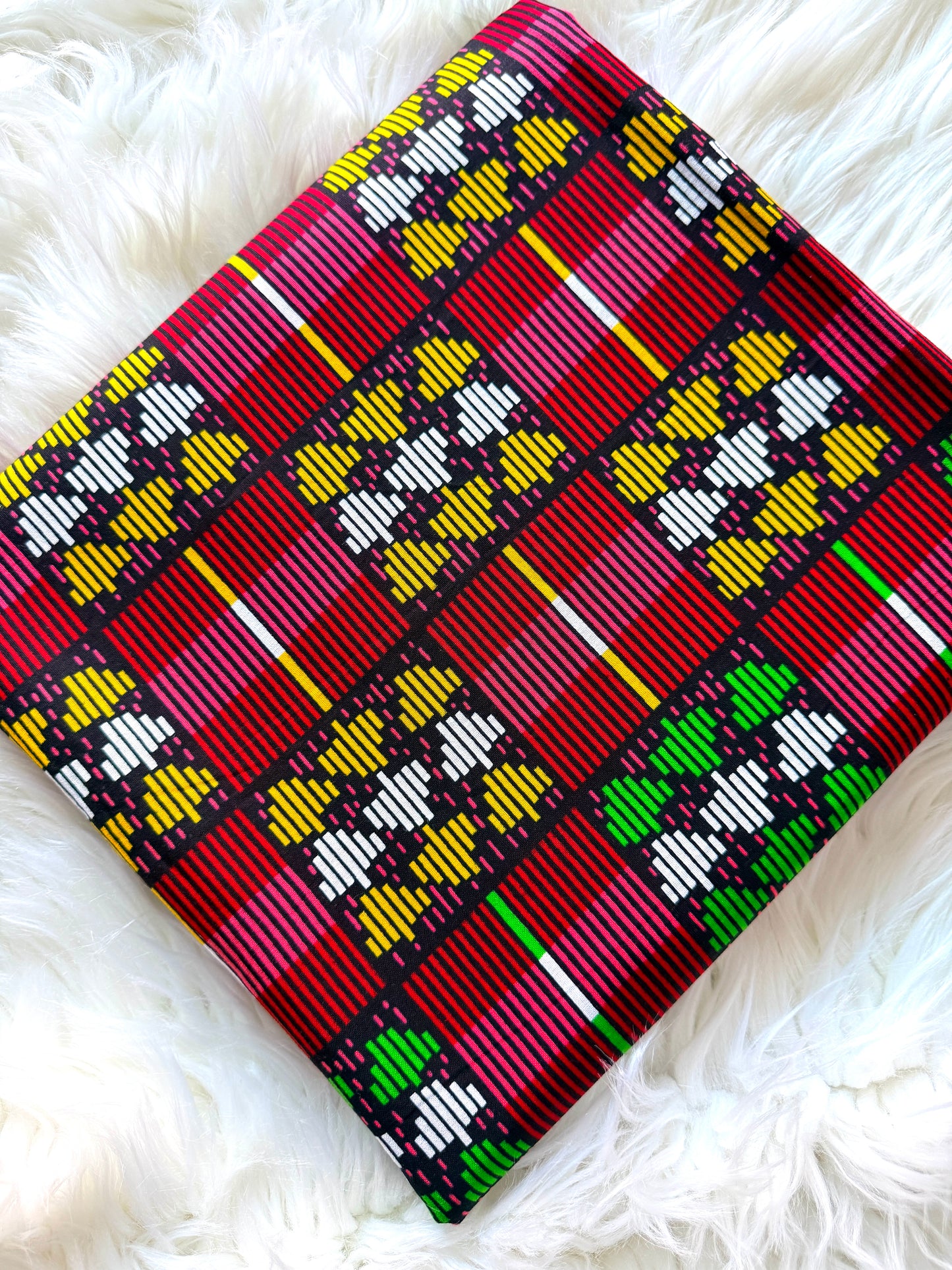 A square woven textile with a bold geometric pattern in red, yellow, green, white, and black, laid flat on a white faux fur surface.