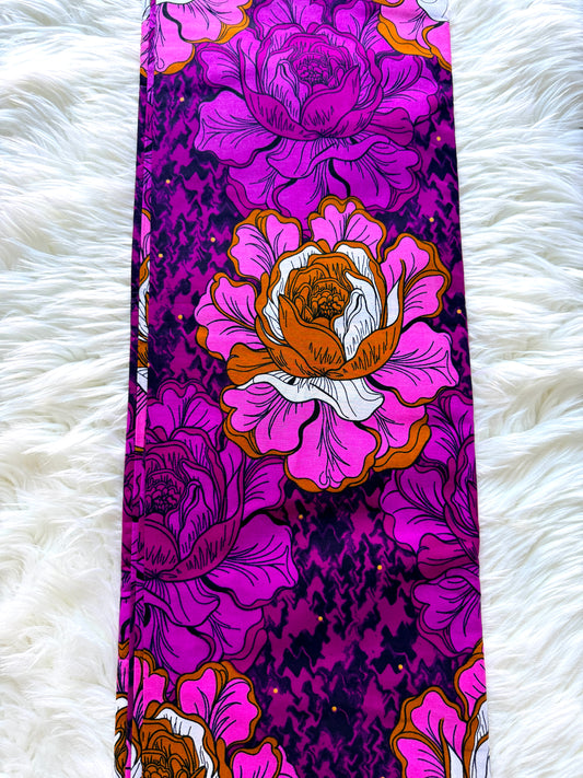 A long, narrow fabric accessory, likely a belt or scarf, featuring a bold floral print with large pink and purple flowers, orange centers, and a dark purple textured background, laid flat on a white faux fur surface.