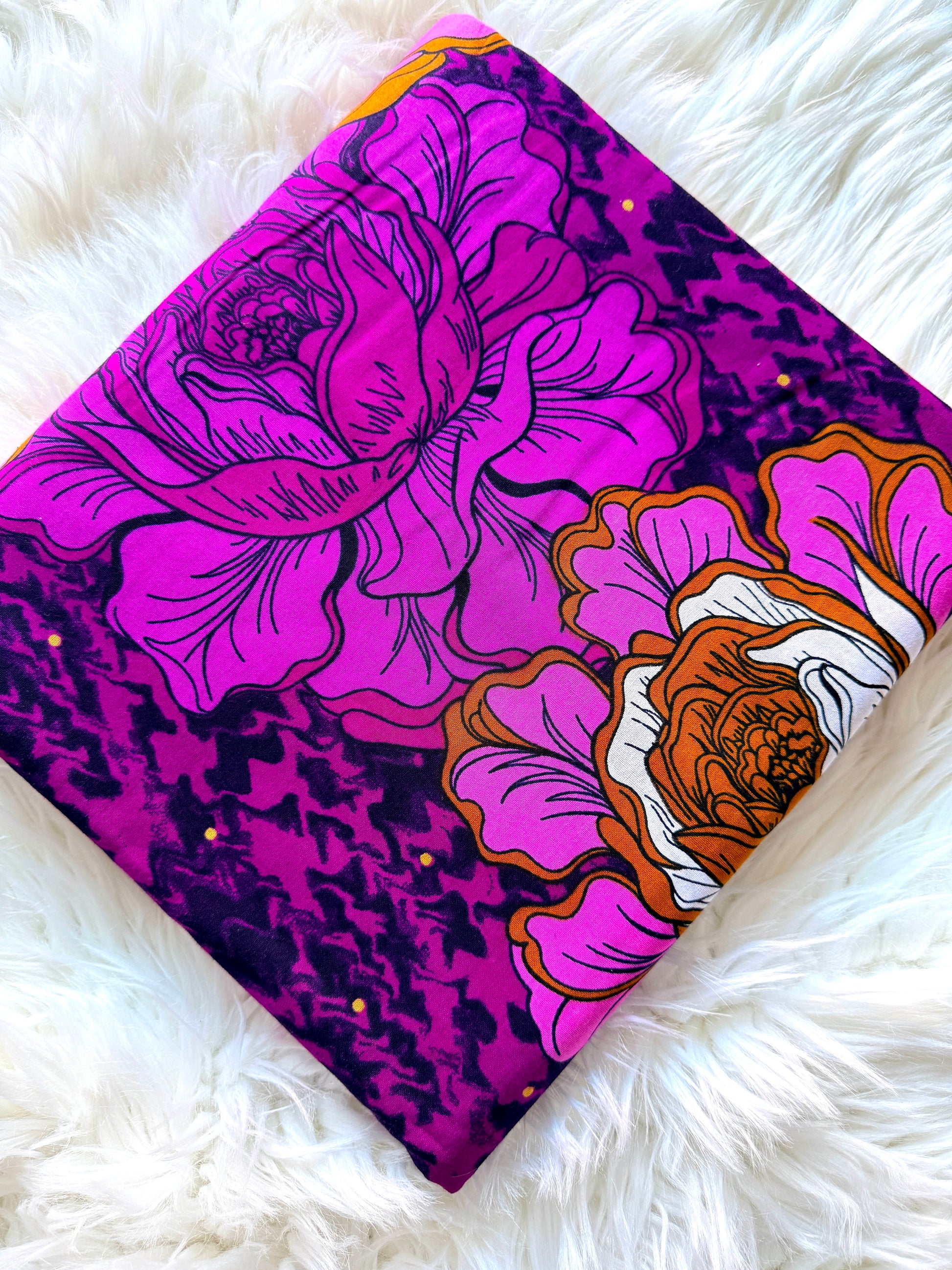 A long, narrow fabric accessory, likely a belt or scarf, featuring a bold floral print with large pink and purple flowers, orange centers, and a dark purple textured background, laid flat on a white faux fur surface.