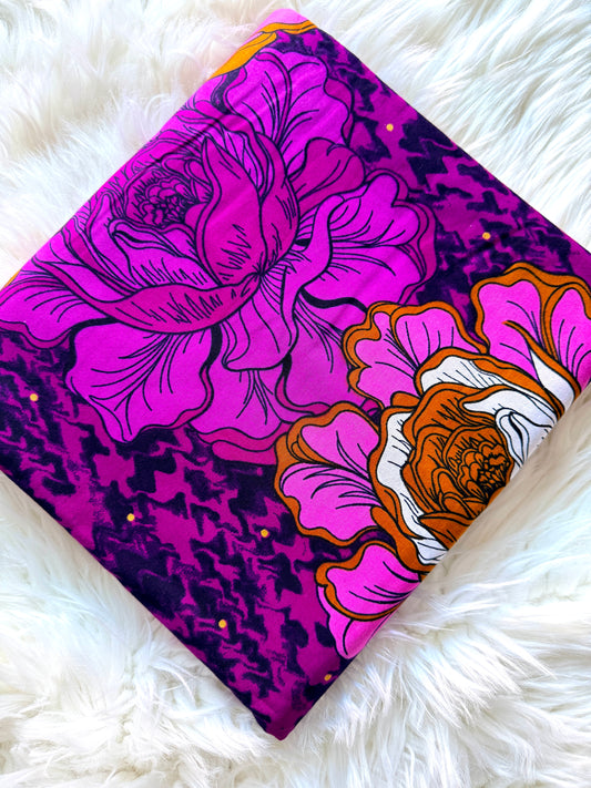 A long, narrow fabric accessory, likely a belt or scarf, featuring a bold floral print with large pink and purple flowers, orange centers, and a dark purple textured background, laid flat on a white faux fur surface.