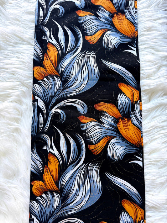A long rectangular scarf laid flat on a white faux-fur surface, featuring a bold abstract floral pattern in orange, light blue, white, and black.