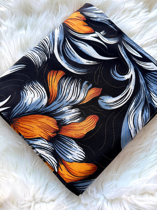 A square fabric item, likely a scarf or small pillow, featuring a bold abstract floral pattern in orange, light blue, and white on a black background, styled on a white faux fur surface.