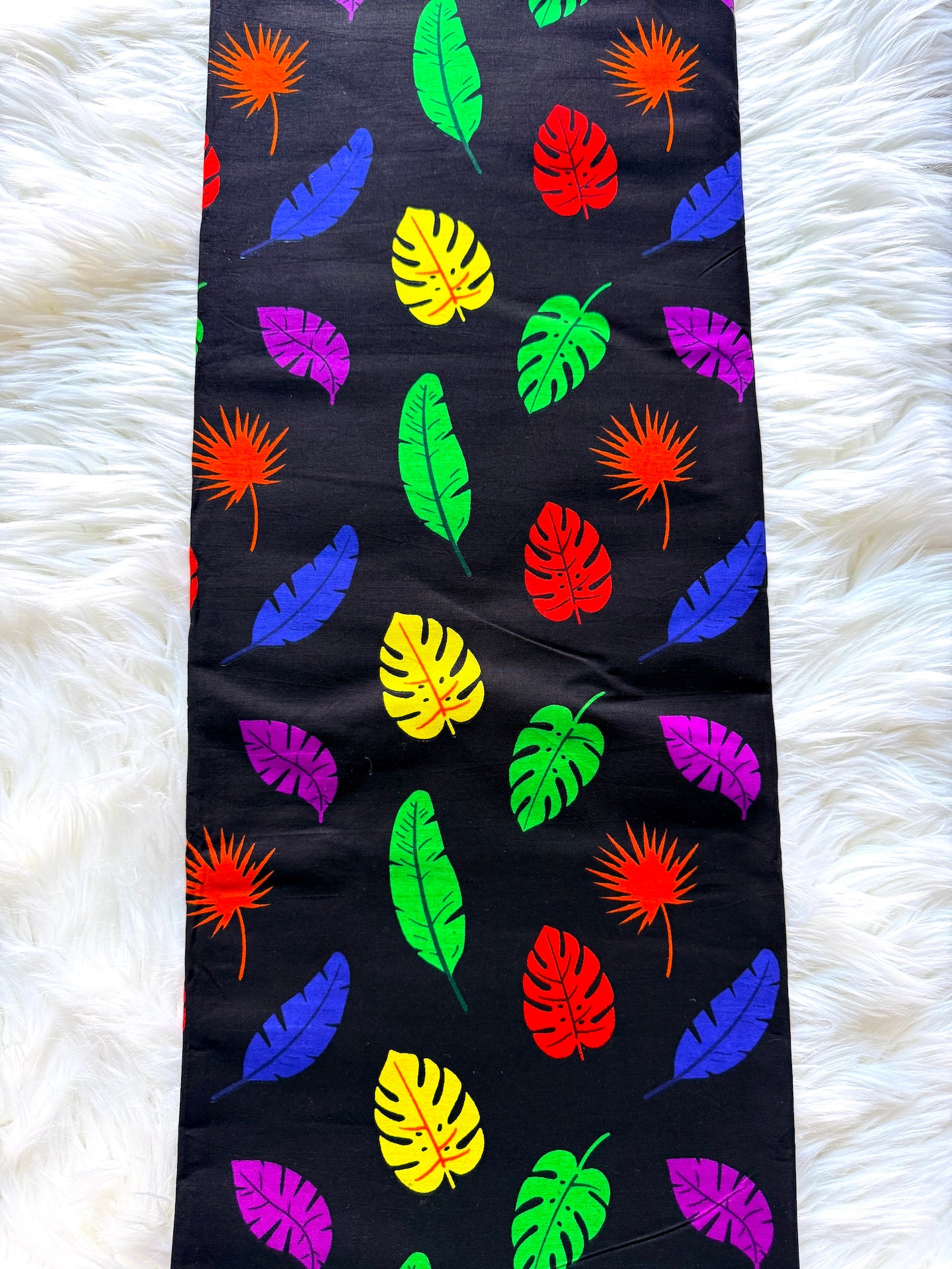 A black fabric table runner or rug featuring a colorful tropical leaf pattern, laid flat on a white faux fur surface.
