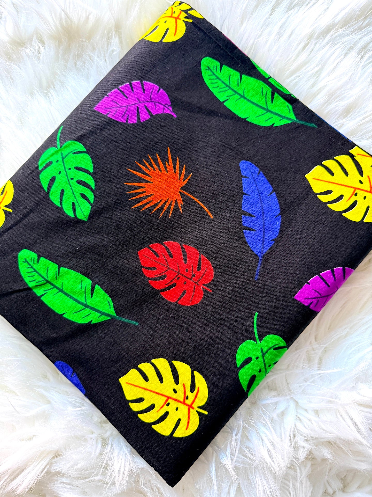 A square fabric item, likely a pillow cover or blanket, featuring a vibrant tropical leaf pattern on a black background, styled flat on a white faux fur surface.