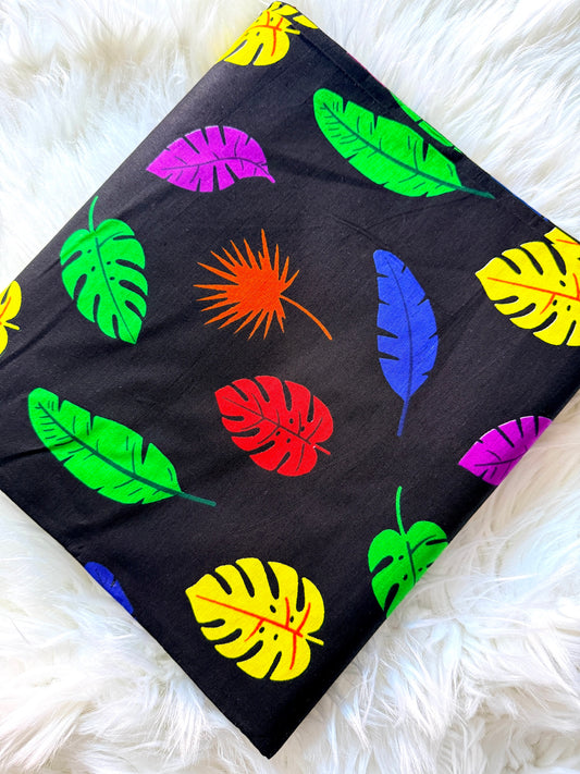 A square fabric item, likely a pillow cover or blanket, featuring a vibrant tropical leaf pattern on a black background, styled flat on a white faux fur surface.