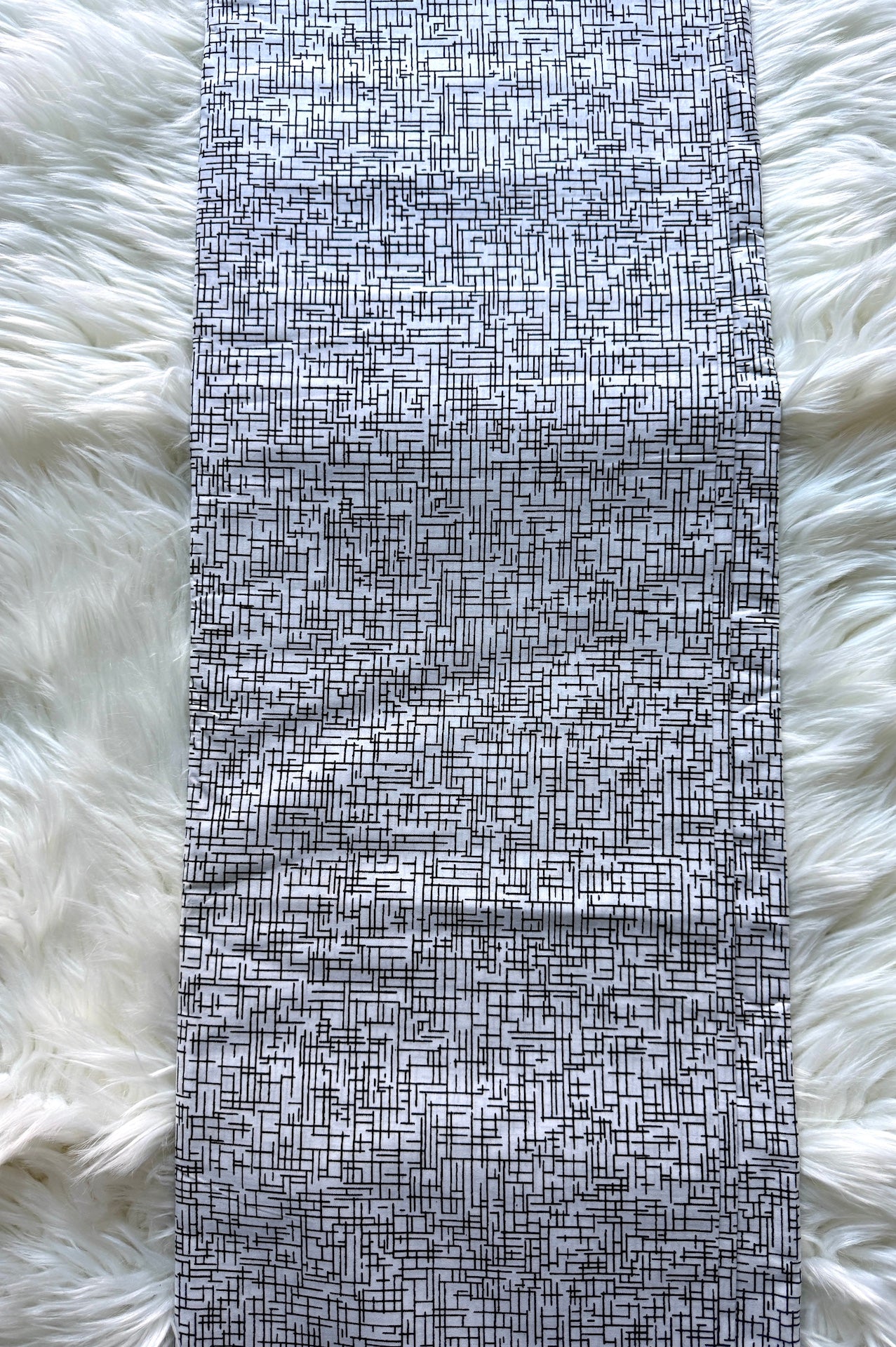 A long rectangular textile item with a black and white abstract linear pattern, laid flat on a white faux-fur surface.