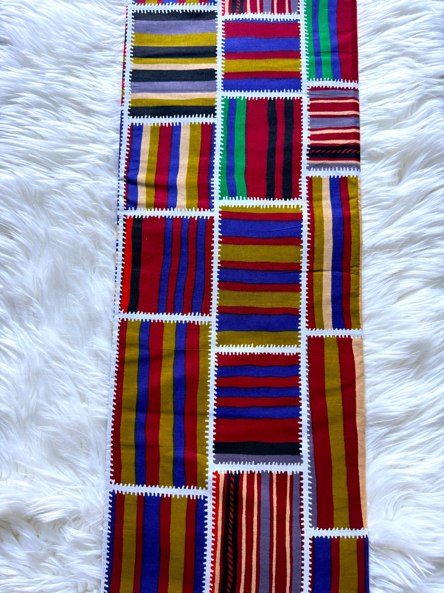 A long woven textile runner with vertical panels of colorful striped patterns in red, blue, yellow, green, black, and white, laid flat on a white faux fur background.