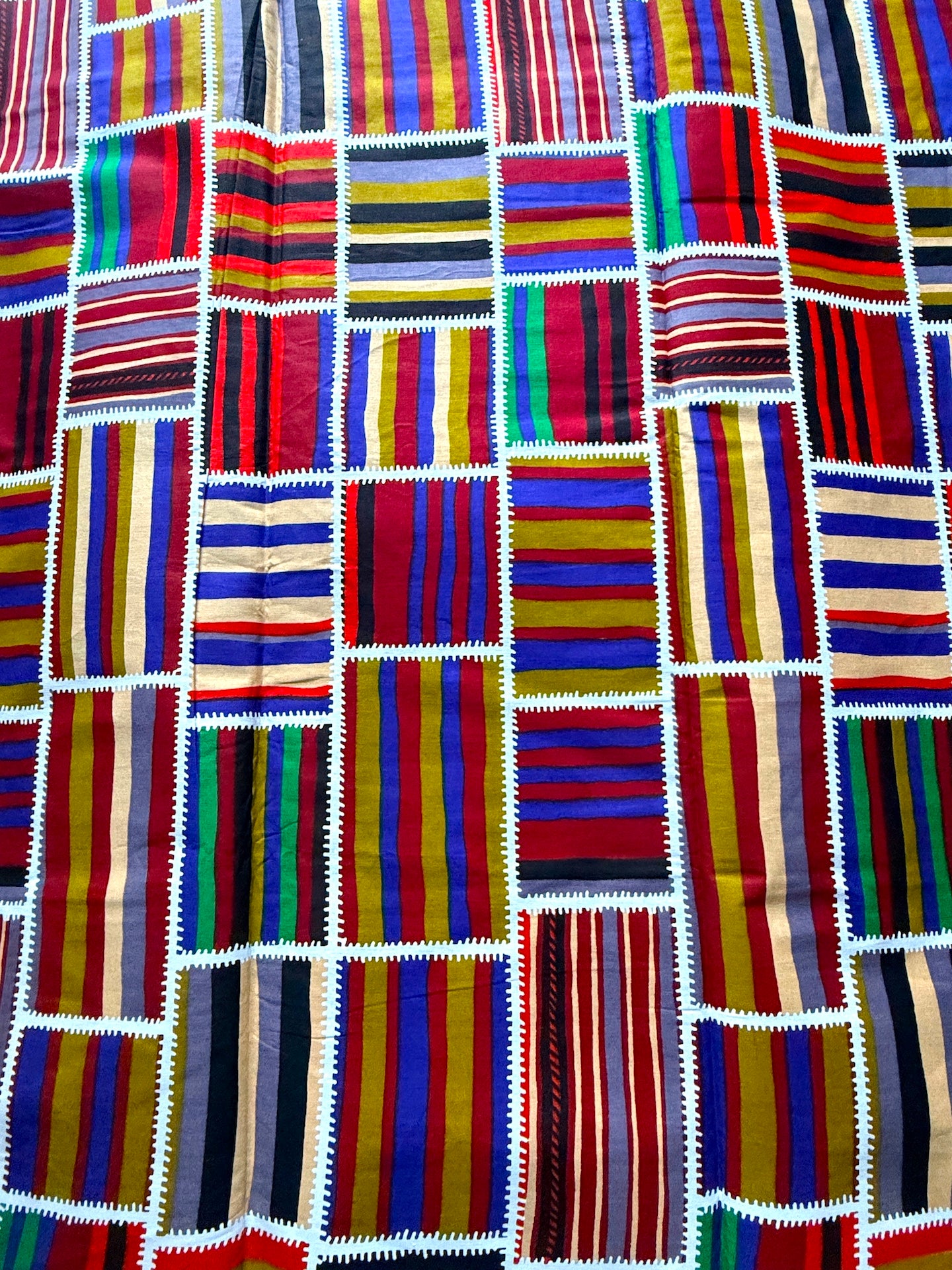 A close-up of a colorful patchwork textile made of many rectangular woven fabric panels with vertical stripes in red, blue, yellow, green, black, white, and tan, stitched together with white zigzag edging.