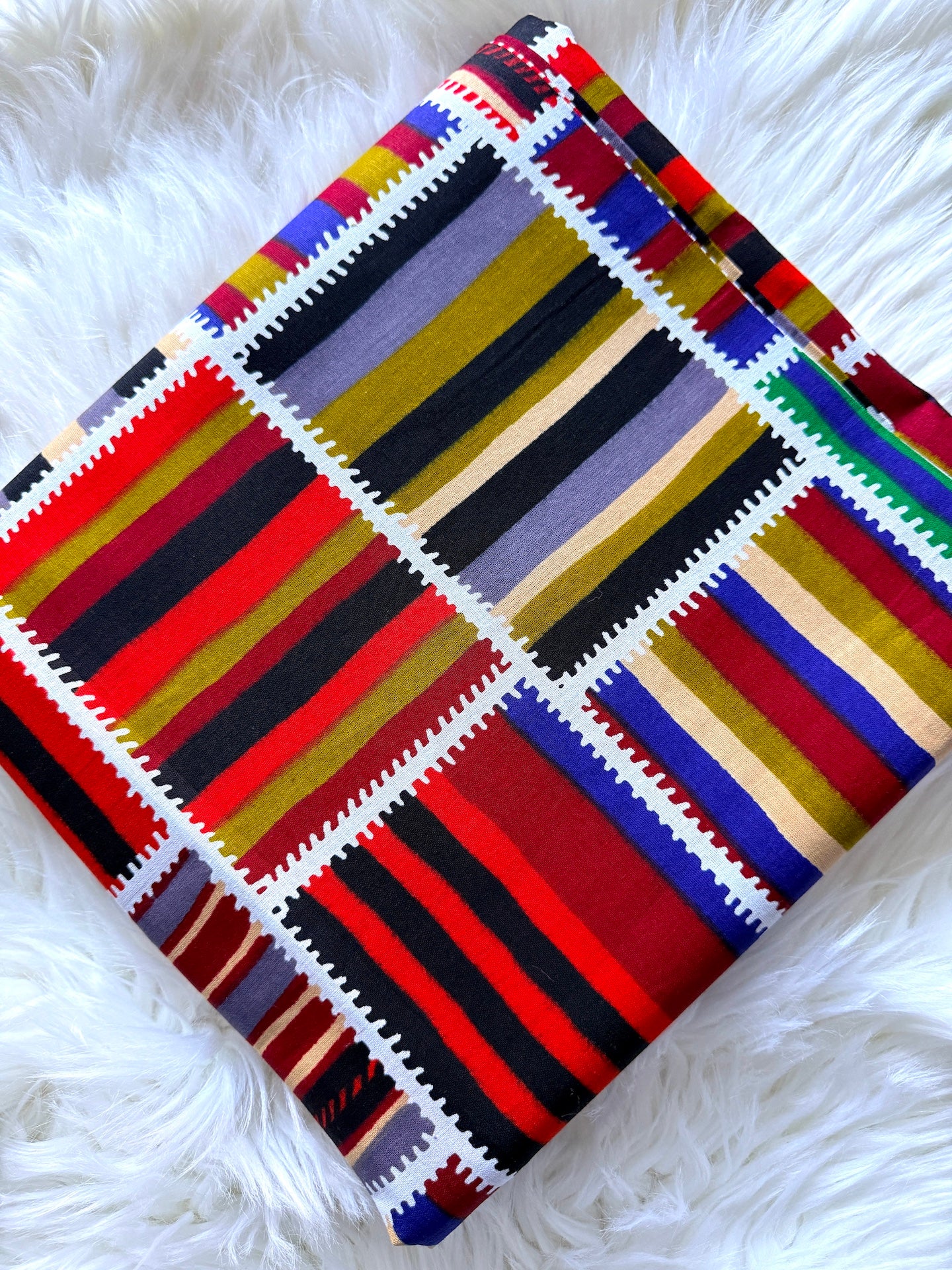 A folded multicolor striped textile, likely a blanket or throw, featuring a patchwork design of red, black, yellow, blue, purple, and beige stripes separated by white zigzag borders, displayed on a white faux fur surface.