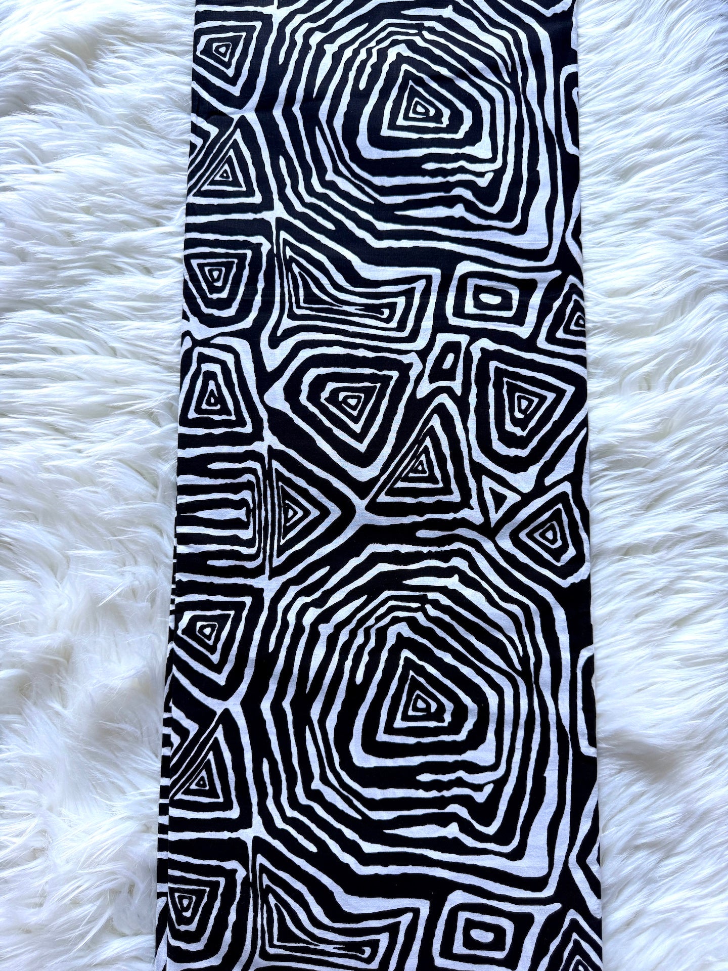 A black and white patterned fabric accessory, likely a scarf or long belt, featuring concentric geometric triangles and zigzags, laid flat on a white faux fur surface.