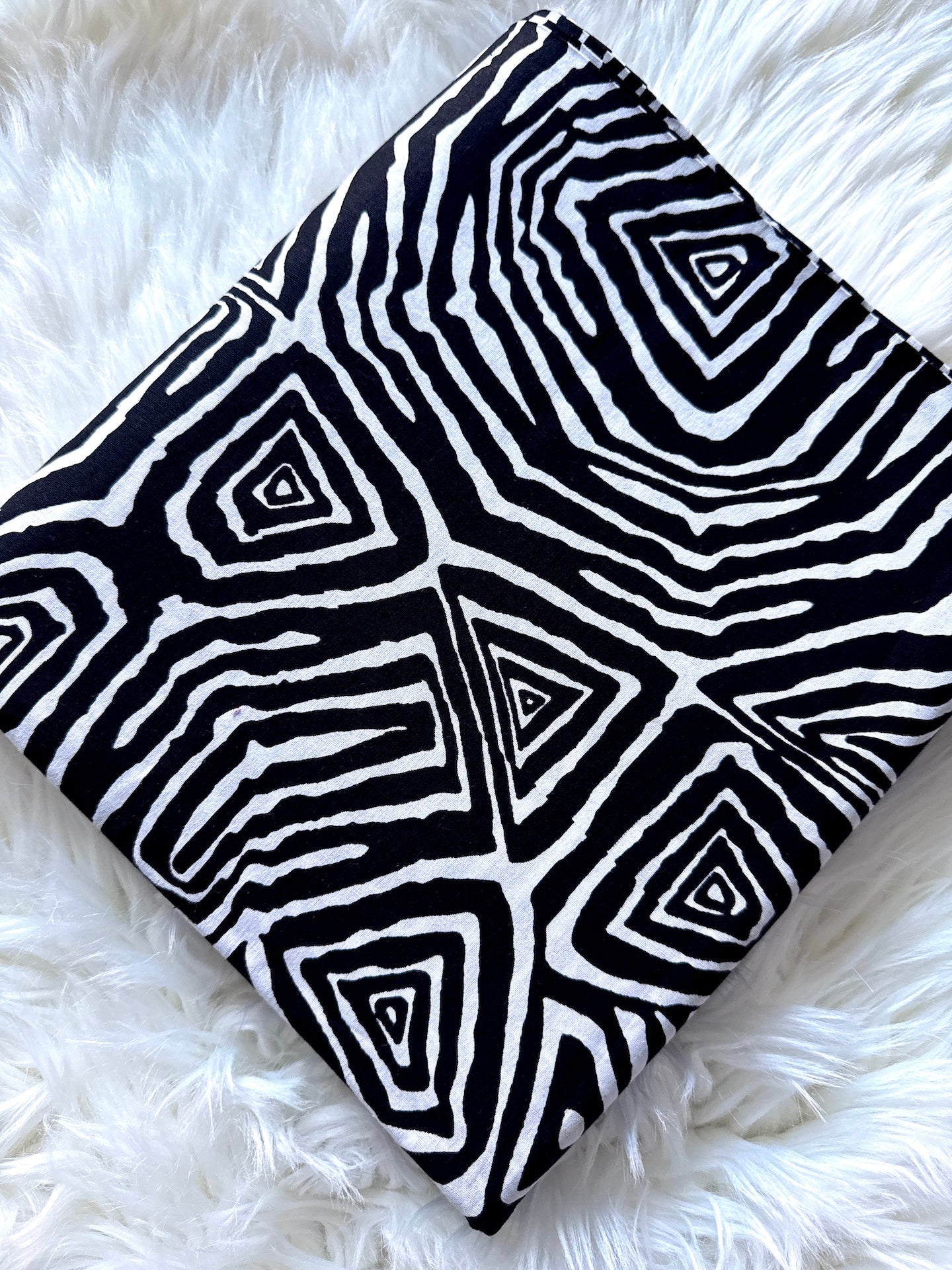 A square fabric pillow or cushion cover featuring a bold black and white geometric pattern with concentric triangles and wavy lines, laid flat on a white faux fur surface.