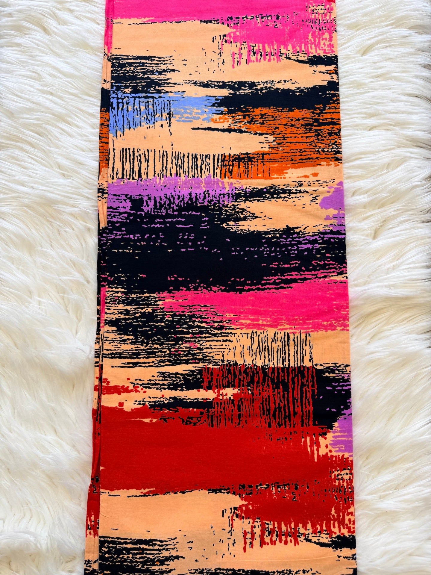 A long rectangular scarf with an abstract brushstroke pattern in vibrant pink, red, orange, purple, black, and beige, laid flat on a white faux fur surface.