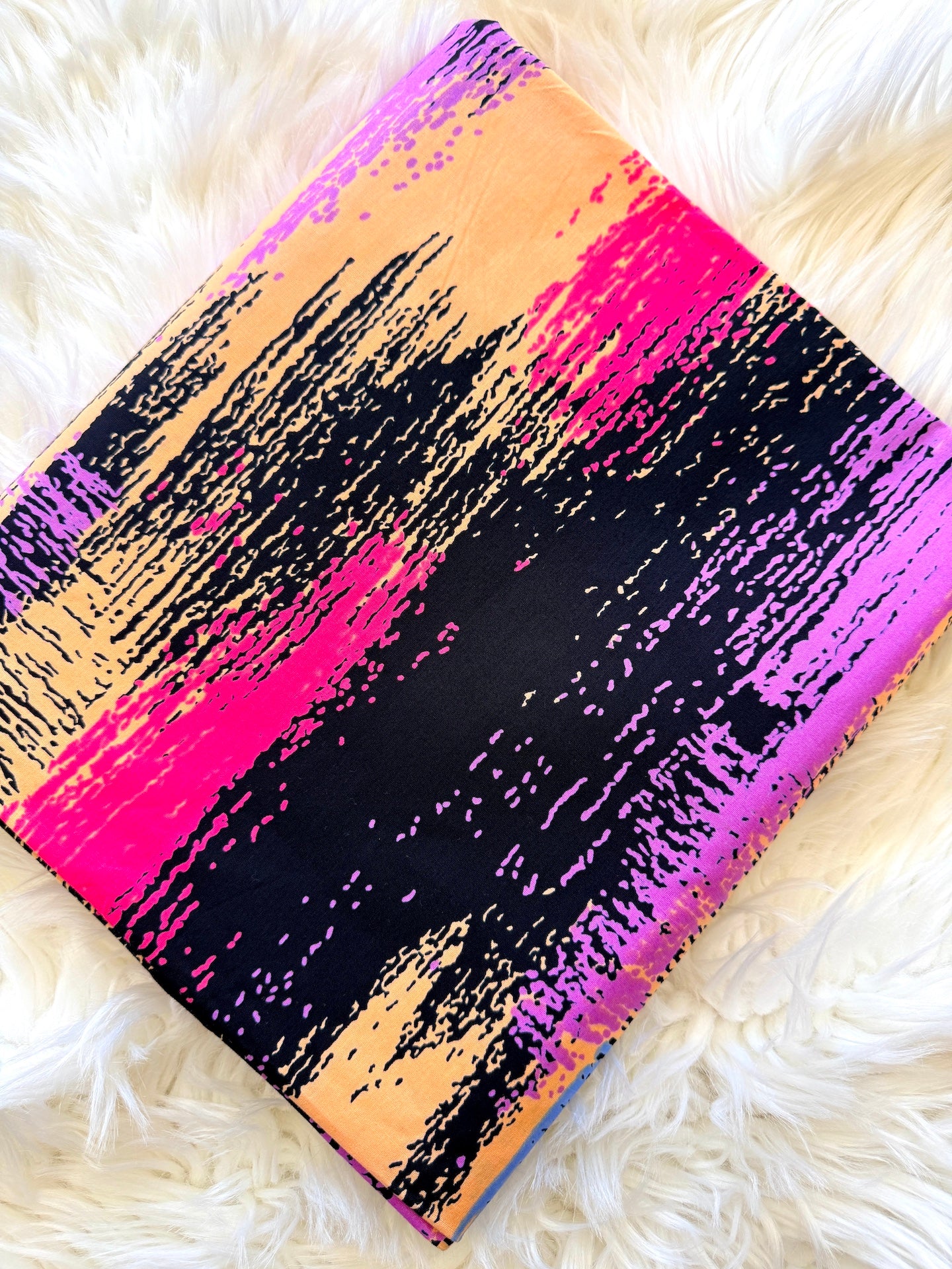 A square fabric item, likely a scarf or small bag, featuring a bold abstract brushstroke pattern in black, hot pink, purple, and peach-orange, laid flat on a white faux fur surface.