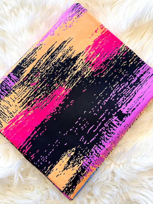 A square fabric item, likely a scarf or small bag, featuring a bold abstract brushstroke pattern in black, hot pink, purple, and peach-orange, laid flat on a white faux fur surface.