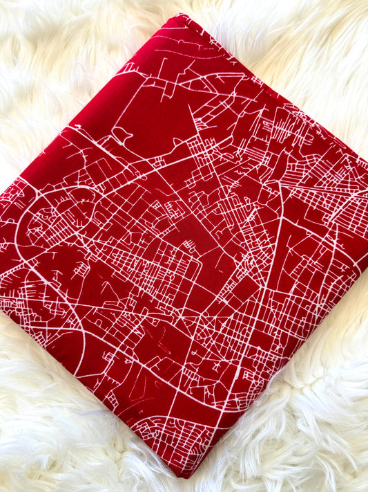 A square red fabric item, likely a pillow cover or small blanket, featuring a white street map pattern, laid flat on a white faux fur surface.