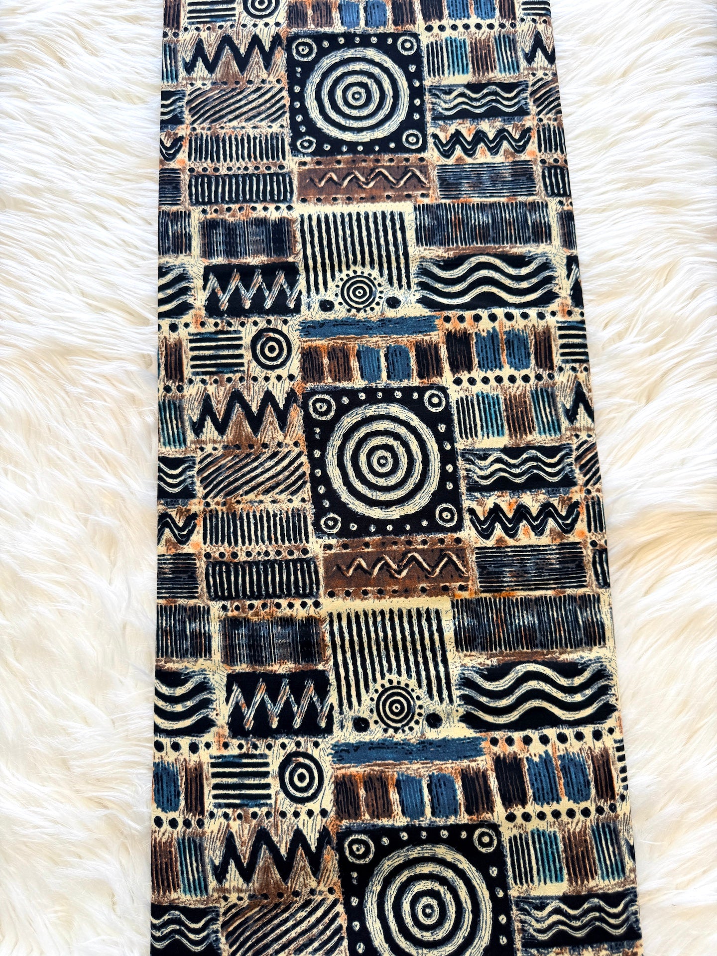 A long rectangular textile runner with an intricate geometric and tribal-inspired pattern, laid flat on a white faux fur surface. The pattern features a mix of black, navy blue, brown, and beige blocks with various abstract designs like spirals, stripes, and wavy lines.