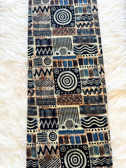 A long rectangular textile runner with an intricate geometric and tribal-inspired pattern, laid flat on a white faux fur surface. The pattern features a mix of black, navy blue, brown, and beige blocks with various abstract designs like spirals, stripes, and wavy lines.