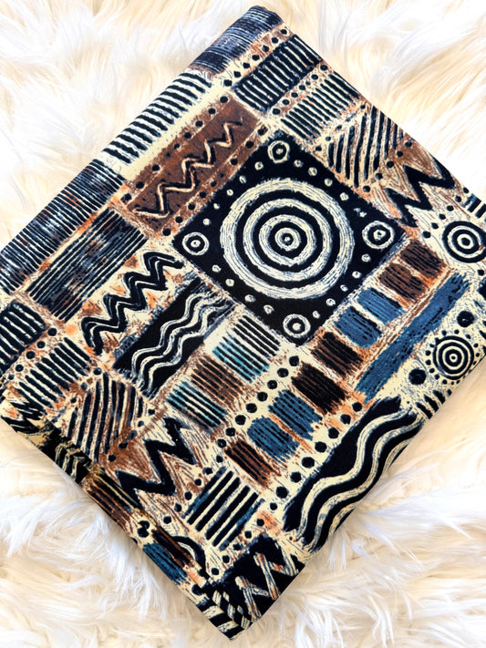 A folded square textile with an intricate patchwork-style geometric and tribal-inspired pattern in black, blue, brown, and cream, displayed on a white faux fur surface.