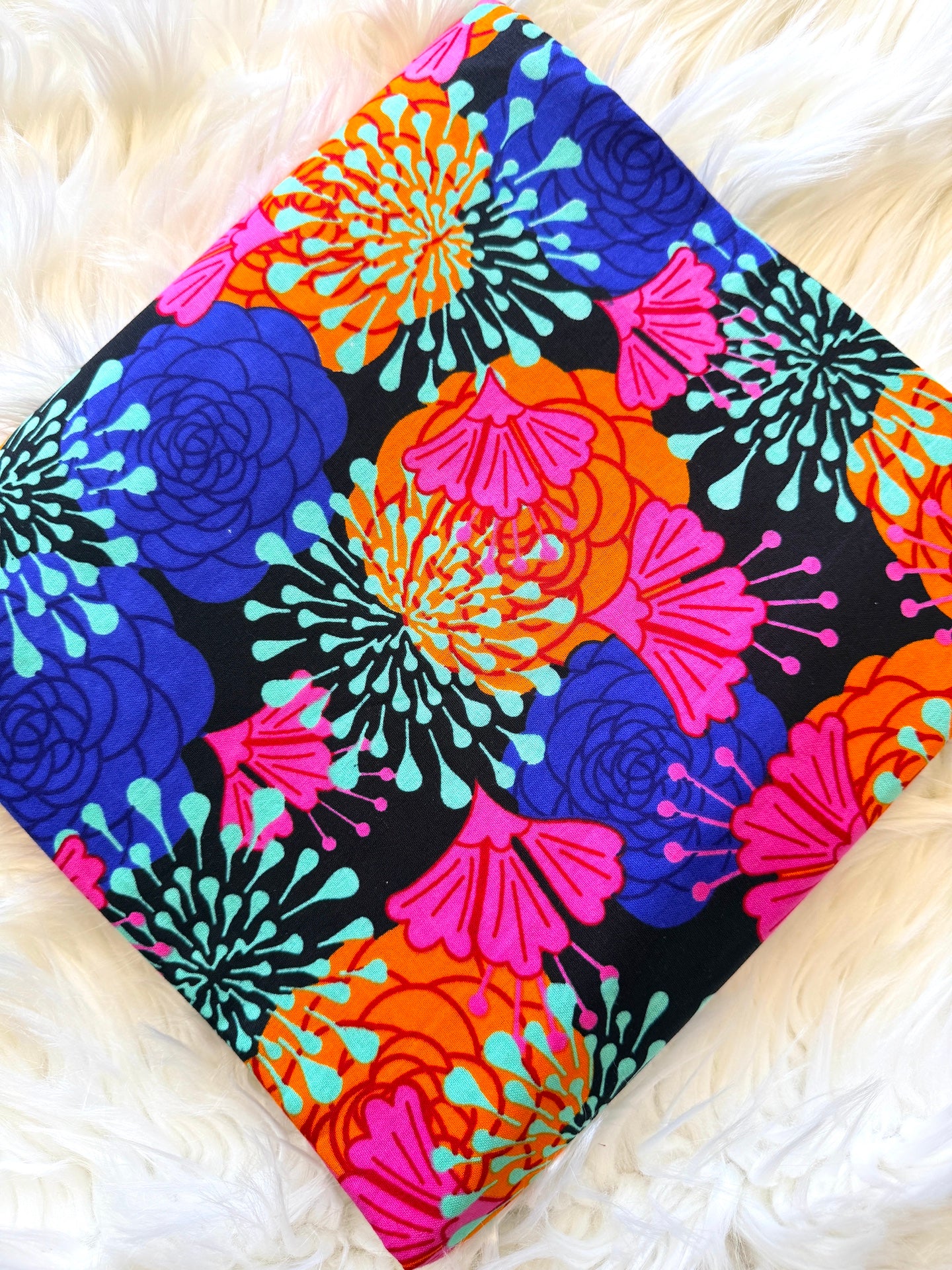 A square fabric item, likely a pillow cover or scarf, featuring a vibrant retro floral pattern on a black background. The design includes bright blue, pink, orange, and turquoise flowers, laid flat on a white faux fur surface.