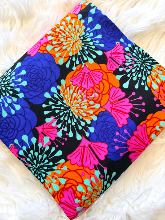 A square fabric item, likely a pillow cover or scarf, featuring a vibrant retro floral pattern on a black background. The design includes bright blue, pink, orange, and turquoise flowers, laid flat on a white faux fur surface.