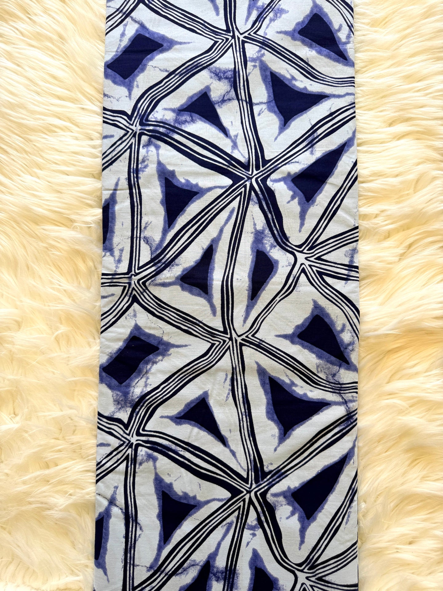 A long rectangular scarf with a blue, white, and black geometric shibori tie-dye pattern, laid flat on a cream-colored faux fur background.