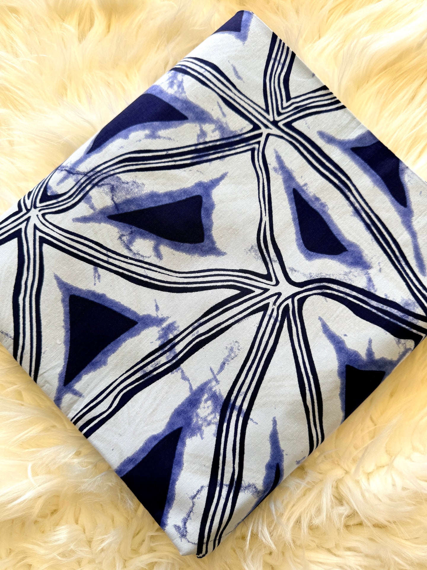 A square textile item, likely a pillow cover or folded blanket, featuring a navy blue and white geometric shibori tie-dye pattern, displayed diagonally on a fluffy cream faux fur surface.