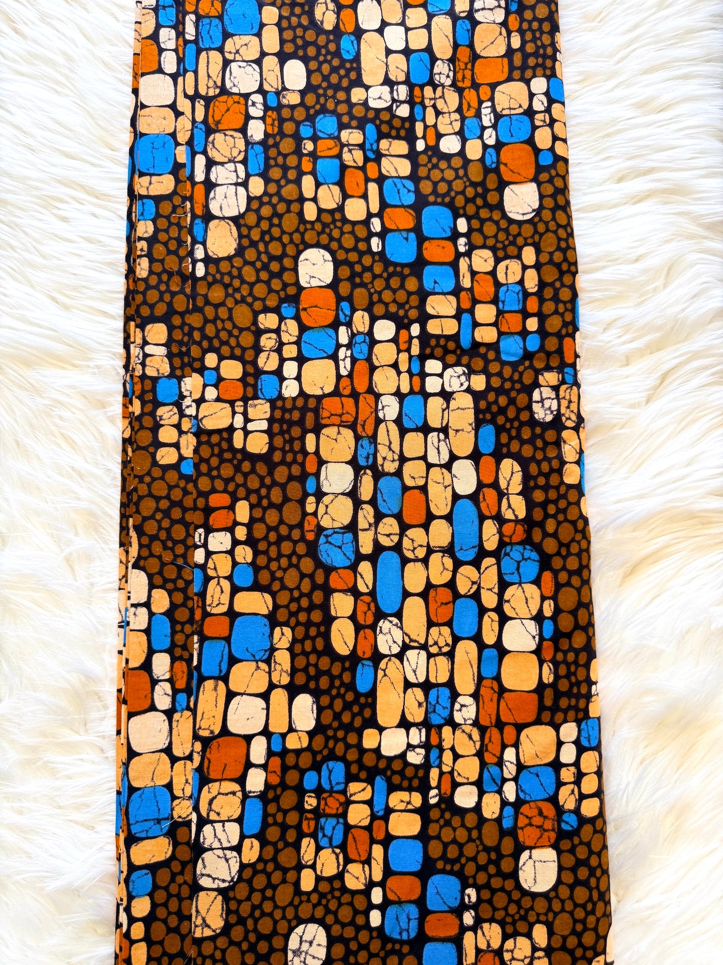 A vertical rectangular textile laid flat on a white faux-fur surface, featuring an abstract mosaic pattern with brown, black, blue, orange, and cream shapes.