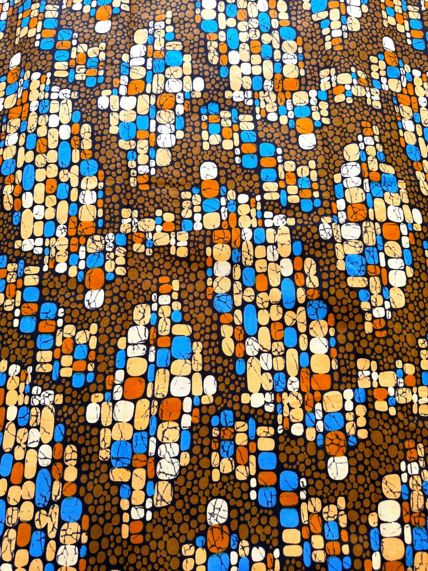 A close-up of a patterned fabric featuring a dense, abstract design of blue, orange, beige, and white rectangular blocks over a dark brown background with small black dots.