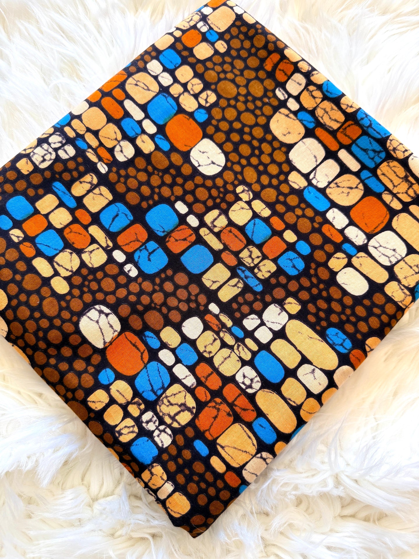 A square piece of African wax print fabric featuring a colorful abstract pattern of stones and dots, laid flat on a white faux fur surface.