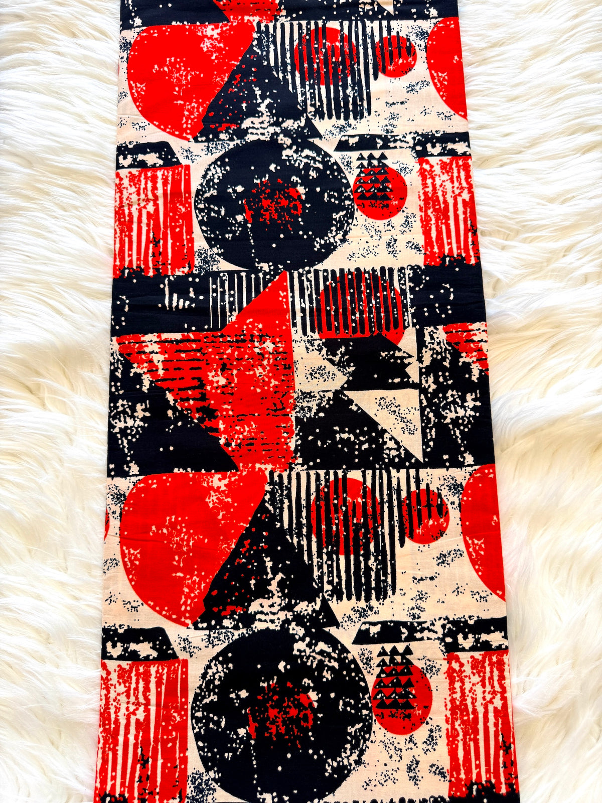 A long rectangular scarf with a bold abstract geometric print in red, black, and cream, laid flat on a white faux fur surface.