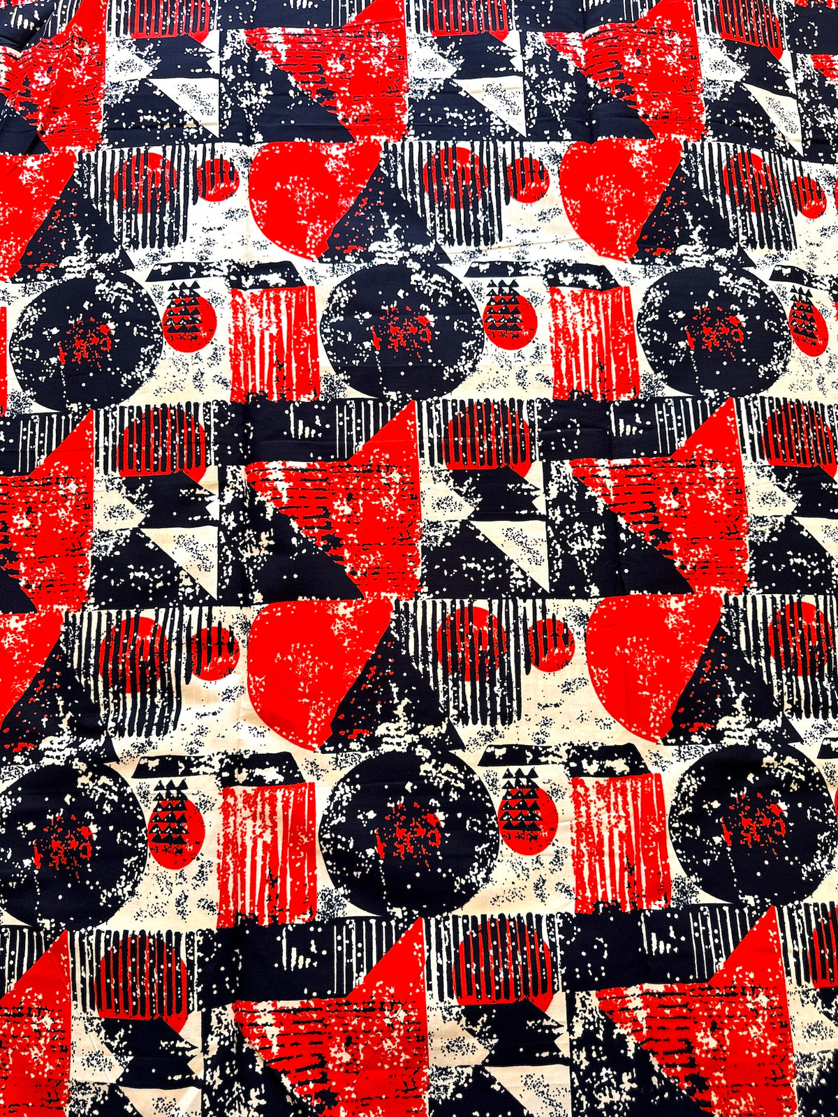 A close-up of a fabric or wallpaper pattern featuring a distressed, vintage-style geometric design in red, black, and off-white. The pattern consists of circles, triangles, and striped blocks.