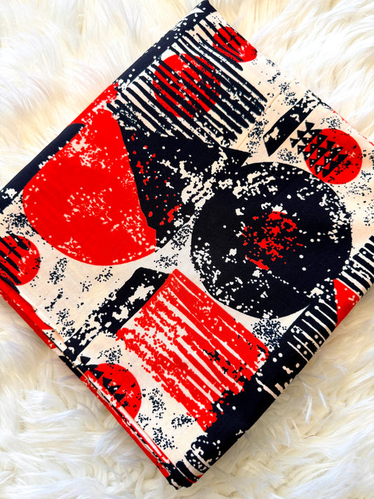 A folded fabric item, likely a blanket or throw, featuring a bold abstract pattern of red, black, and cream with distressed textures, laid flat on a white faux fur surface.