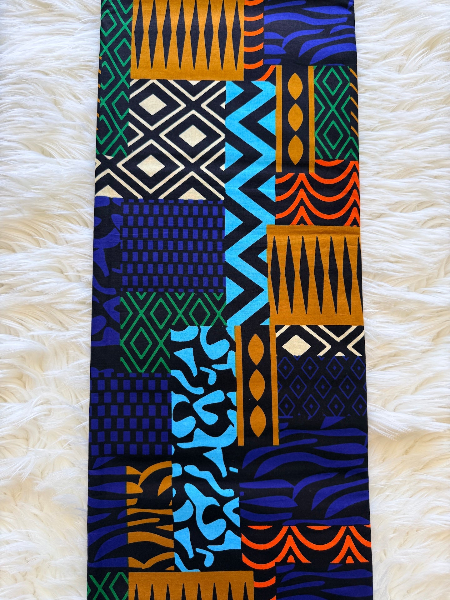 A long rectangular textile laid flat on a white faux-fur surface, featuring a bold multicolor geometric and tribal-inspired patchwork pattern with blue, orange, green, black, and cream sections.