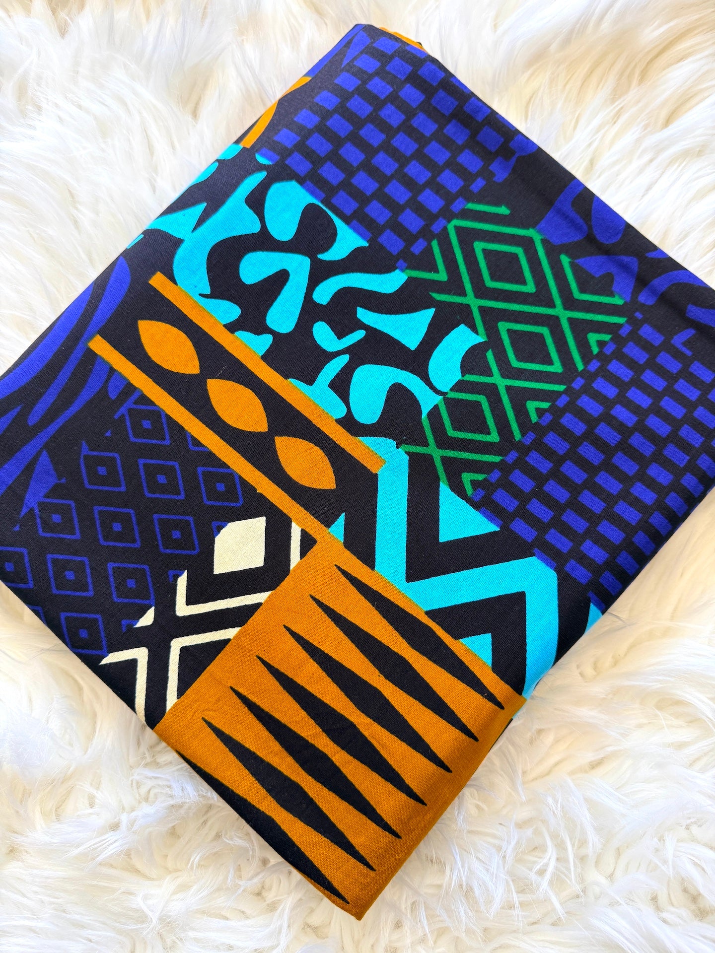 A folded piece of African wax print fabric with a vibrant geometric pattern in blue, black, orange, and green, displayed on a white faux fur background.