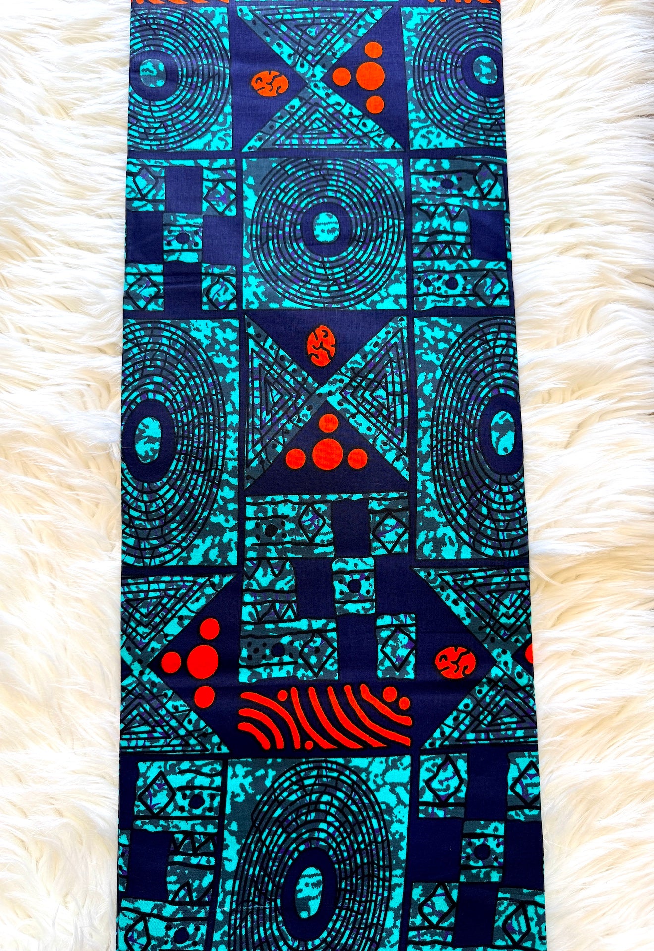 A rectangular fabric panel with a bold geometric and tribal-inspired print in turquoise, black, and red, laid flat on a white faux fur background.