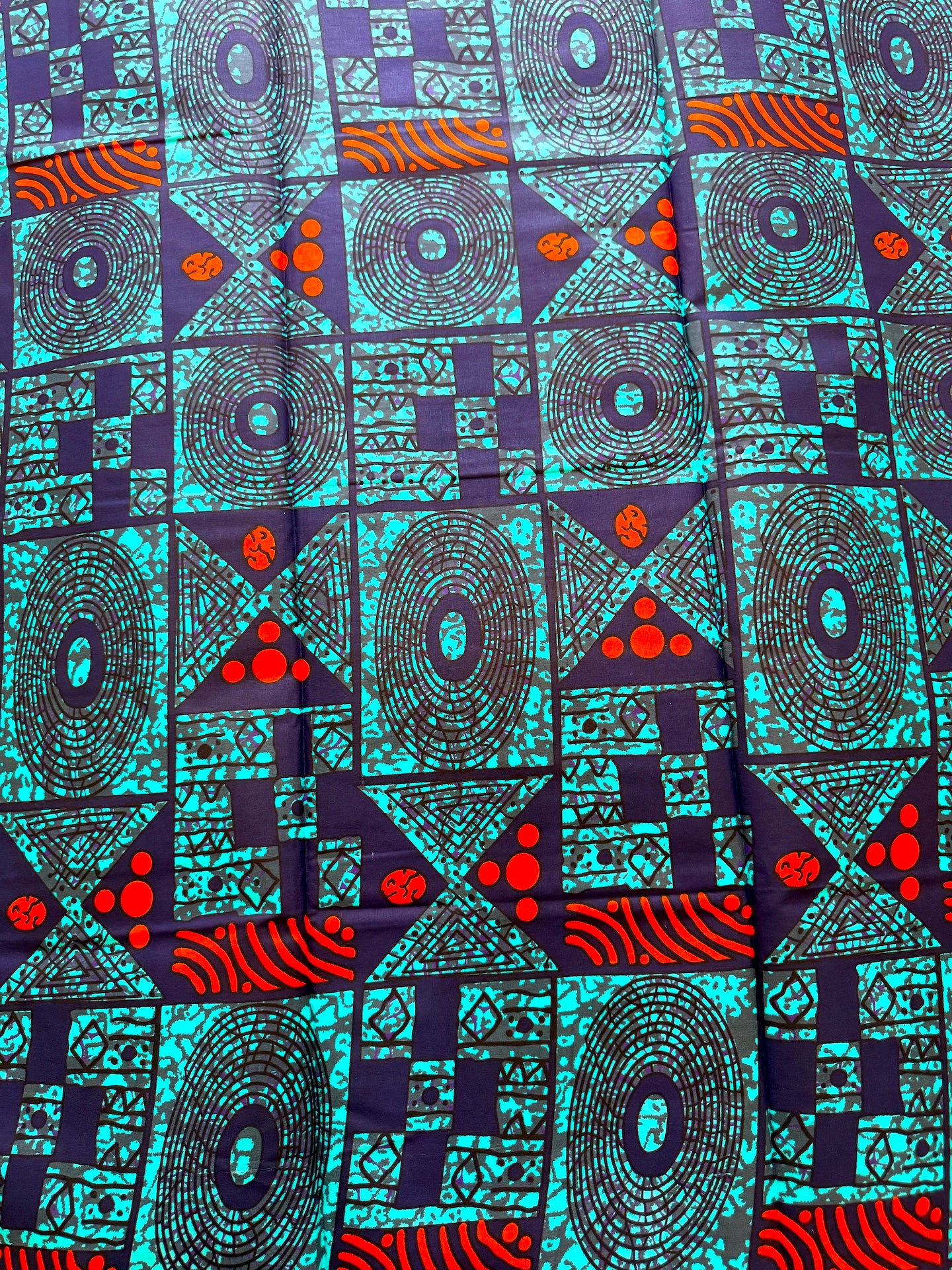 A close-up of a vibrant African wax print fabric featuring geometric patterns in turquoise, dark purple, black, and bright red-orange.