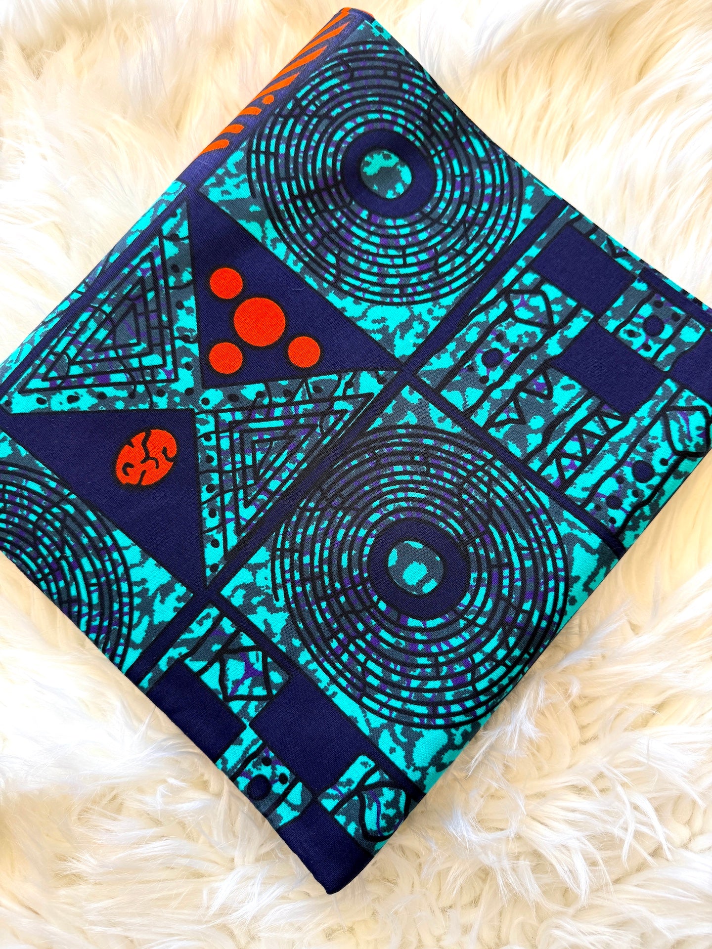 A square fabric item, likely a pillow cover or small blanket, featuring a bold geometric and tribal-inspired print in turquoise, navy, and black, with small orange circular accents, laid flat on a white faux fur surface.