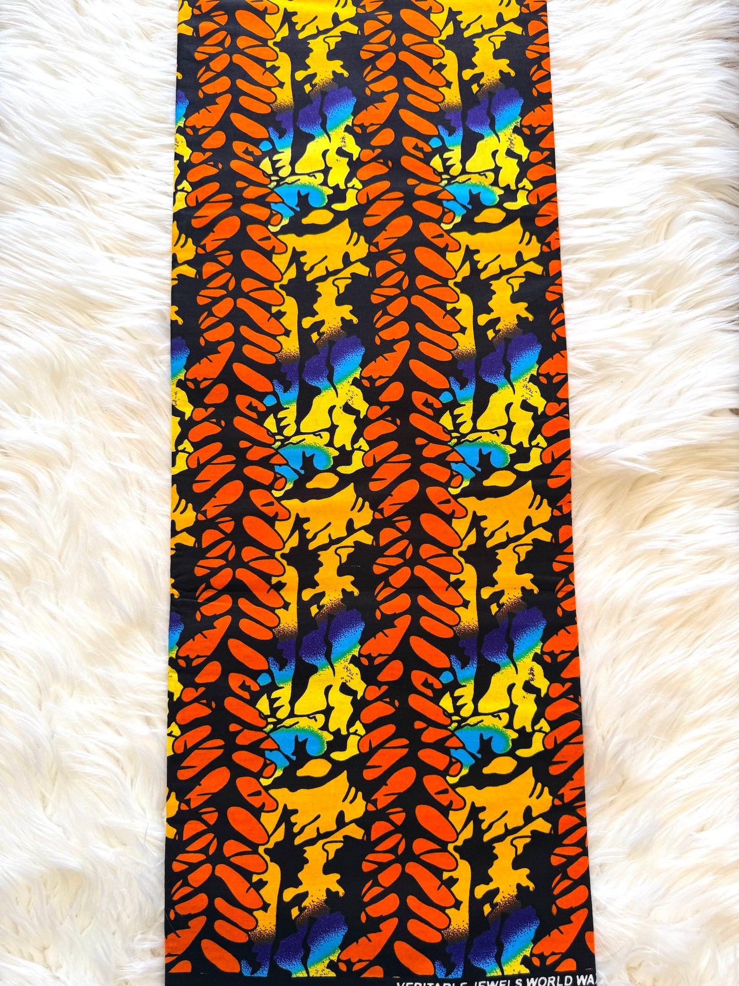 A long rectangular textile laid flat on a white faux-fur surface, featuring a bold vertical abstract pattern in orange, yellow, blue, black, and white.