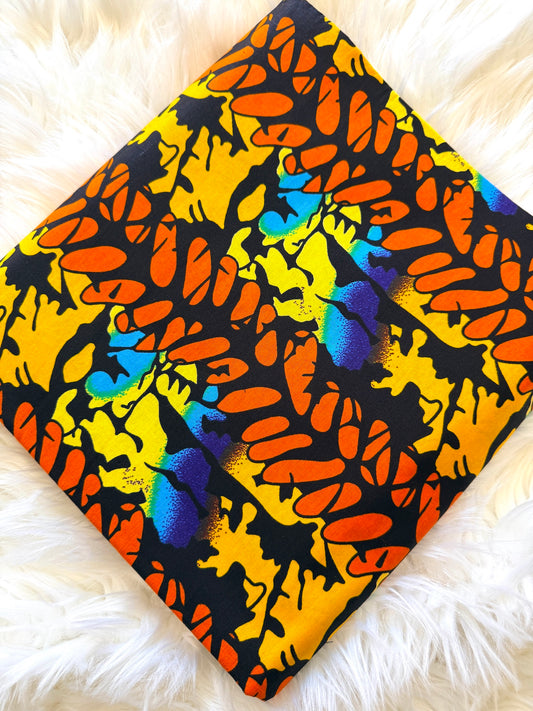 A square fabric item, likely a pillow cover or scarf, featuring a bold abstract pattern in orange, yellow, black, blue, and purple, laid flat on a white faux fur surface.