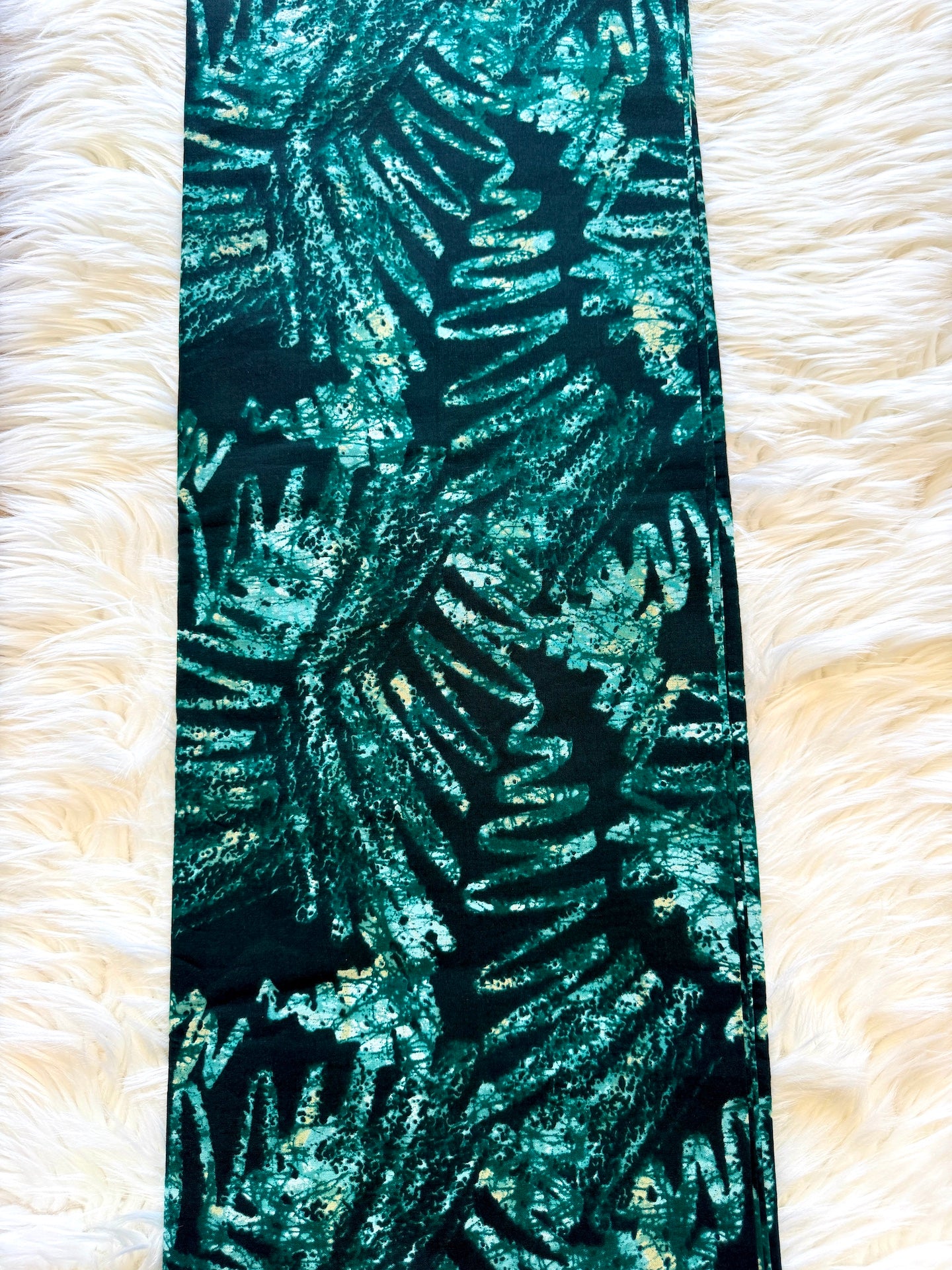A long rectangular fabric panel with a metallic teal and black tropical leaf pattern, laid flat on a white faux fur background.